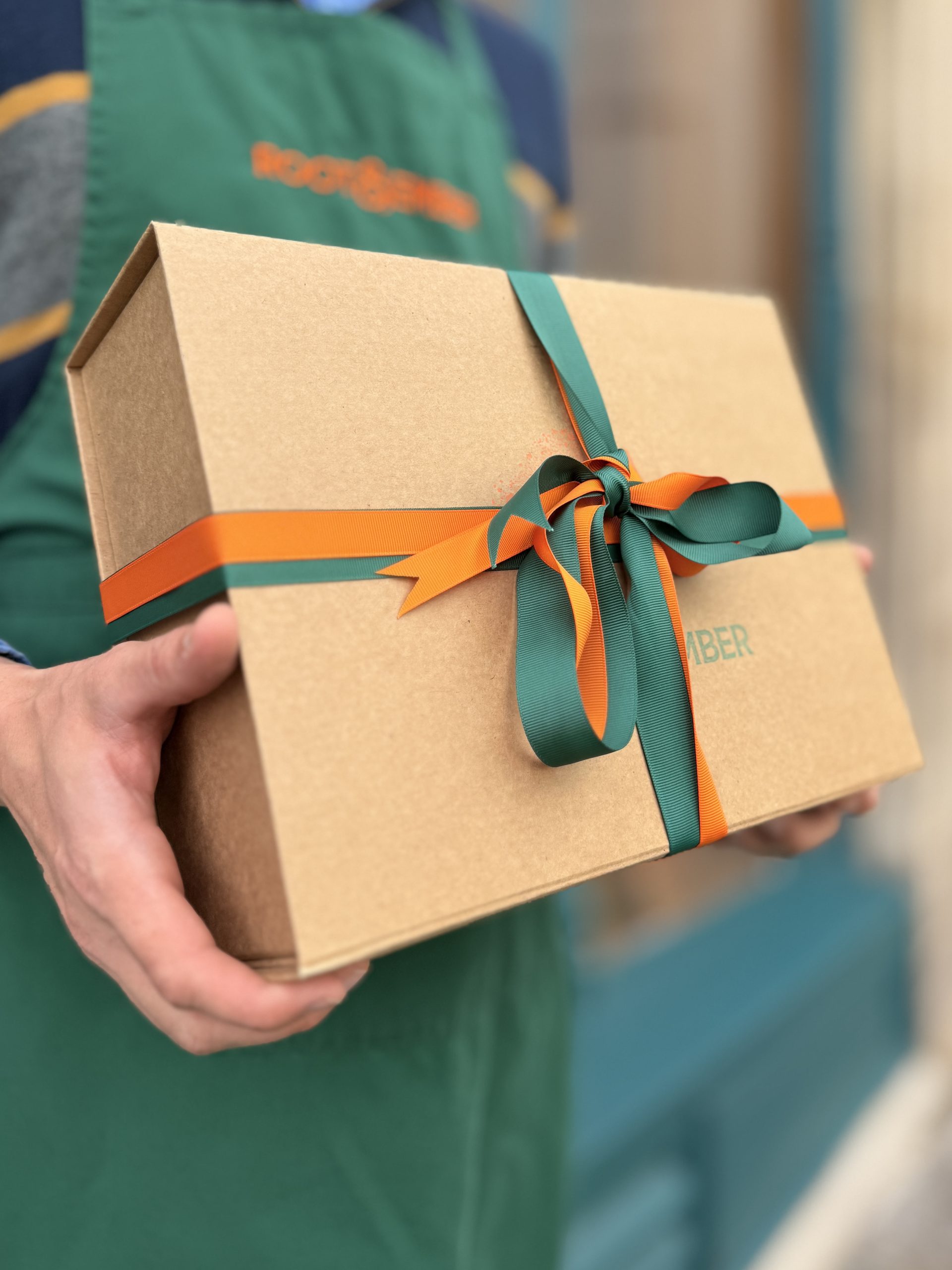 A gift box tied with ribbon, held at the shop door by a member of the team in a green apron